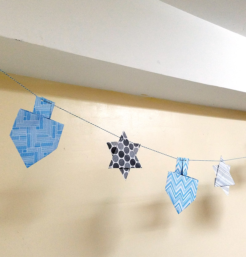 Make a fun origami Hanukkah garland featuring folded paper dreidels and star of david. This simple Chanukah craft for kids teens and adults is a perfect way to update your Hanukkah decor.
