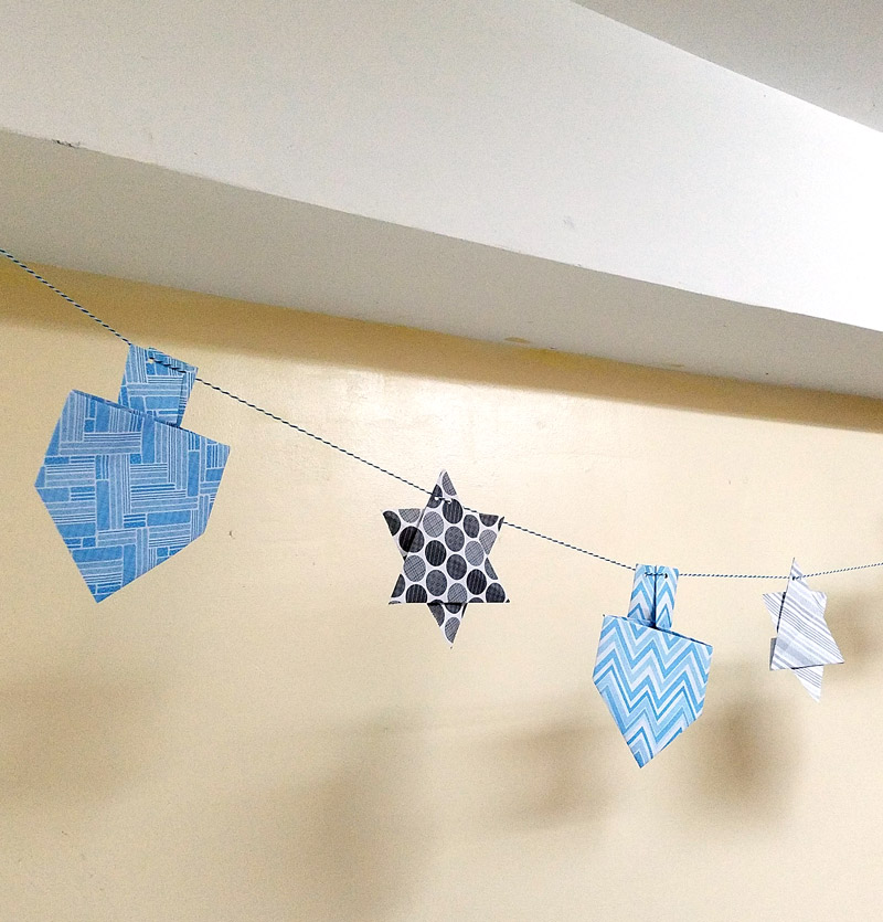 Make a fun origami Hanukkah garland featuring folded paper dreidels and star of david. This simple Chanukah craft for kids teens and adults is a perfect way to update your Hanukkah decor.