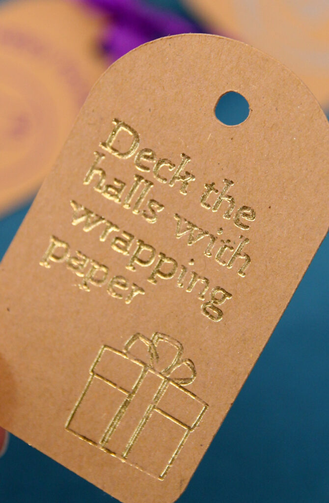 an image of a brown kraft board cricut gift tag with a gold debossed gift box and text that says "deck the halls with wrapping paper". 